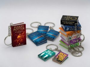 Assorted miniature book keychains on metal rings and chains, featuring a red Harry Potter book, blue Game of Thrones miniatures, a stack of The Lord of the Rings books, and separated titles like The Hobbit, Half Girlfriend, and Rich Dad Poor Dad on a white background.