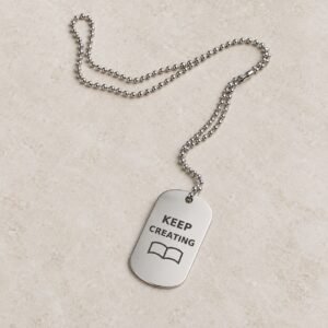 Keep Creating author dog-tag necklace with dark words and an open-book motif on a glossy silver tag.